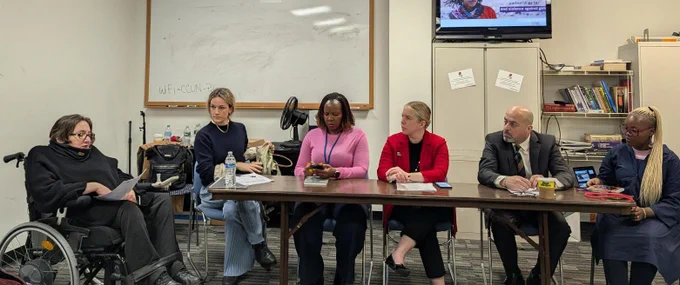 This image shows a group of people gathered around a table in a professional setting. Ola Abualghaib is speaking.
In the background, there is a whiteboard with a note about WiFi and a bookshelf, indicating a workspace or meeting room. Additionally, a screen in the upper right corner displays an image related to gender-based violence.