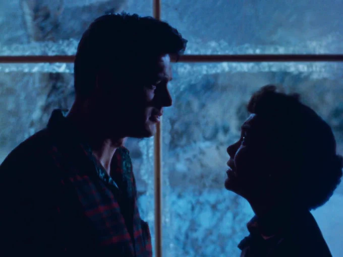 A man and a woman look at each other, silhouetted against a blue window.