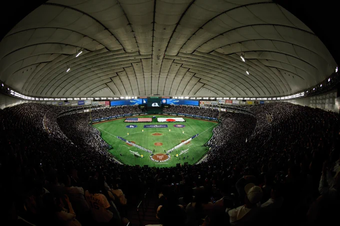 A wide-angle shot of inside the Tokyo Dome during Game 1 of the Tokyo Series between the Dodgers and Cubs. The American flag, Japanese flag and team logos can be seen on the field.