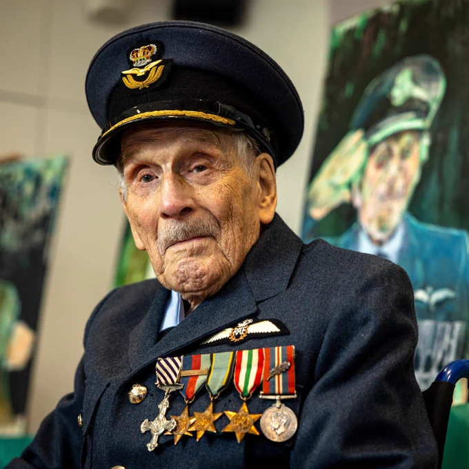 John 'Paddy' Hemingway DFC, dressed in a formal Royal Air Force uniform adorned with five medals and decorations, sits in a wheelchair. He wears a dark blue dress hat with a gold Royal Air Force emblem. Behind him, two portraits are displayed.