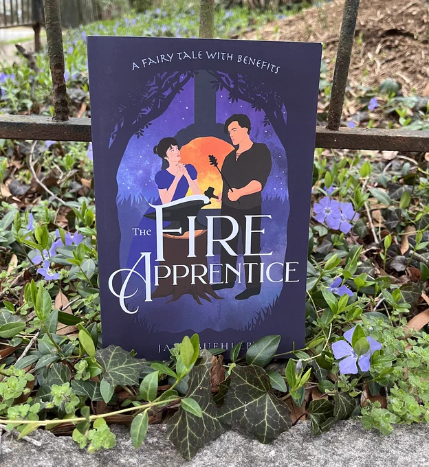 The book The Fire Apprentice showing a woman leaning on an anvil and a man holding a hammer and iron rod, both in front of a glowing hearth under the trees; the book is leaning on an iron railing and resting on top of ivy and blooming blue vinca flowers
