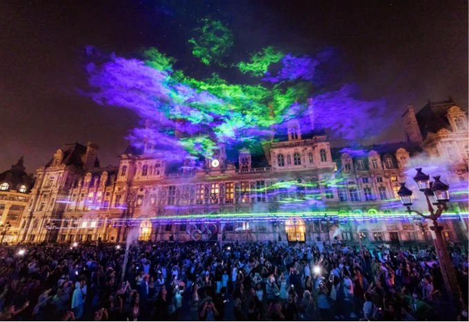 A vibrant light show projected onto the facade of the Hôtel de Ville in Paris, with a large crowd gathered to watch the colorful display in the evening.