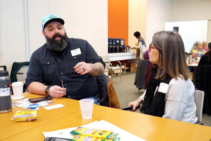 A man with a beard wearing a teal cap and black shirt engaged in conversation with a woman with glasses at a table. Conference materials and refreshments are visible on the table, suggesting a networking or break session.