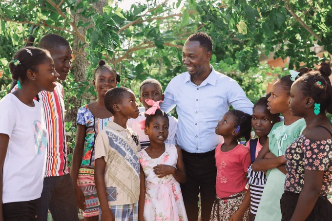 Group of children and an adult interacting happily outdoors with trees in the background.