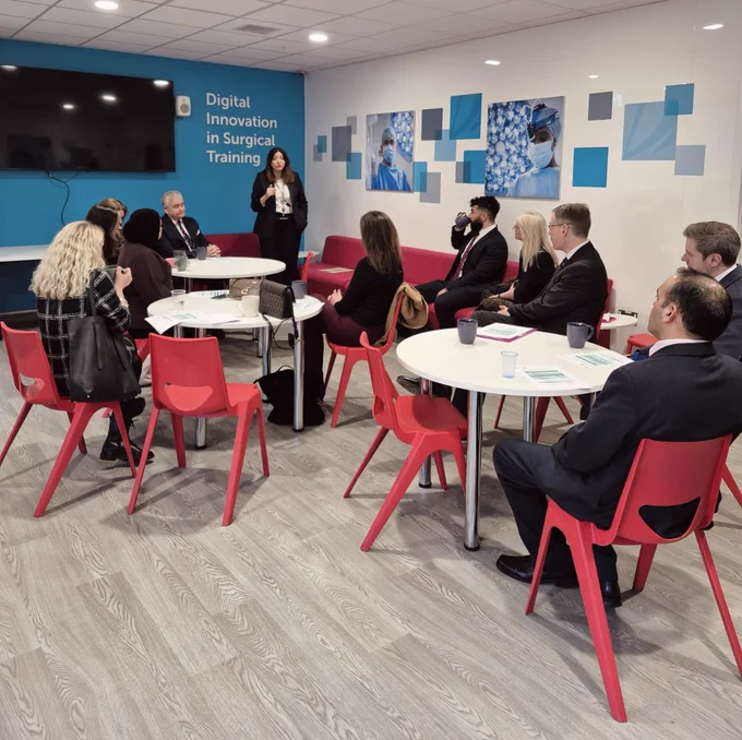 People sit around tables listening to a woman speak. Behind her is a sign which reads 'Digital innovation in Surgical Training'