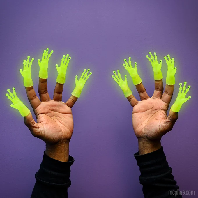 Two hands facing palms out stick out from the bottom of the screen. Every finger has a glowing knobby, green alien hand finger puppet on the tip. The background is purple. 
