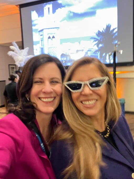 Two individuals smiling at a conference with a projected image of a scenic view in the background. One person is wearing star-shaped sunglasses and a gold necklace, the other is dressed in a bright pink blazer.