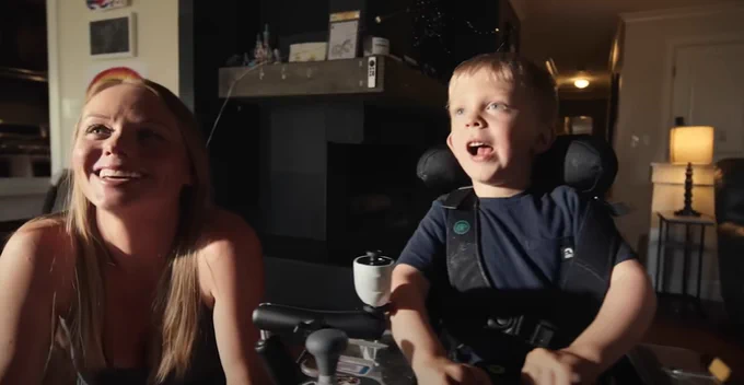 A child uses an adaptive joystick while a family member looks on, smiling. 