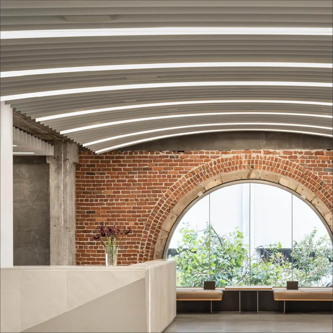 Interior view of a modern office space featuring a blend of exposed brick archways, concrete columns, and curving white ceiling stripes, complemented by minimalistic furniture and potted greenery.