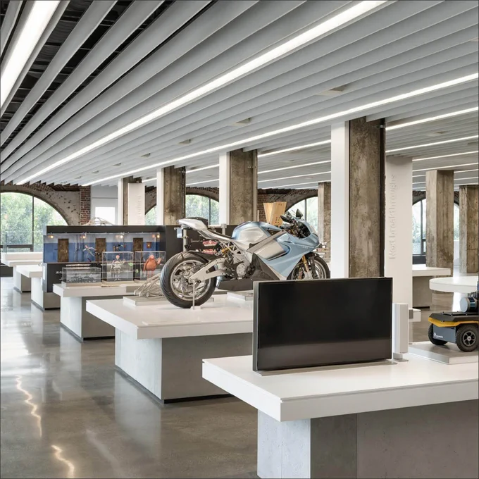 Interior view of a modern museum display featuring a variety of exhibits including a prominent motorcycle, interactive screens, and model cars. The design incorporates sleek, clean lines with natural and artificial lighting.