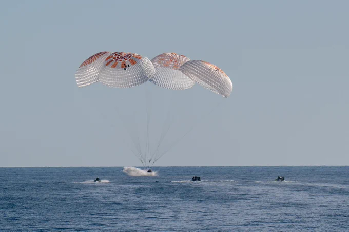 SpaceX's Dragon capsule, a small cone, splashes down into the water off the coast of Florida. Its four main parachutes billow above it, dominating the image, with Dragon and a few small support ships below. The water is fairly calm and the sky is a clear, pale blue. Credit: NASA/Keegan Barber