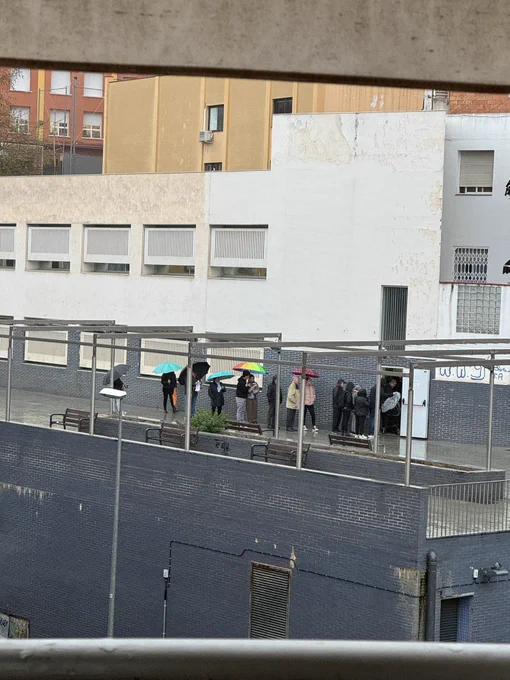 Cola de gente, esperando en la calle, bajo la lluvia. Algunos, no llevan paraguas.