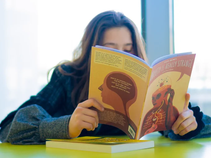 Photo of a teenage girl sat at a table reading a book called 'Anxiety is really strange'.