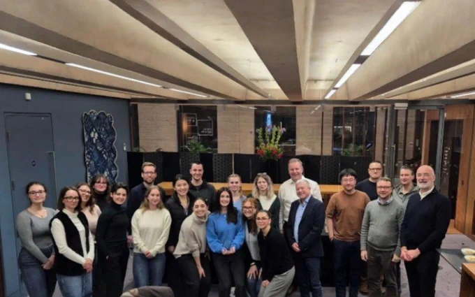 A group of 24 students and staff from UCL and Triad, of mixed genders and diverse clothing, pose for a photo before the hackathon event. They are standing indoors in a well-lit room with wood and glass decor, arranged in three rows, and smiling at the camera