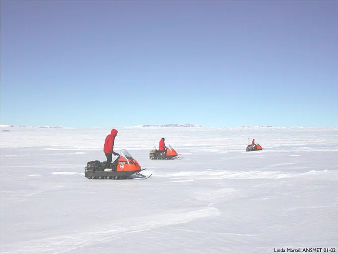 Three researchers in red jackets are riding orange snowmobiles across a vast, snow-covered landscape in Antarctica. The sky is clear and blue, with distant low hills visible on the horizon. One researcher is standing on a stationary snowmobile in the foreground, while the other two are further away, traveling in the same direction. The snow reflects the bright sunlight, and the terrain appears flat and expansive. The image credit in the bottom right corner indicates it was taken by Linda Martel during the ANSMET 01-02 expedition.