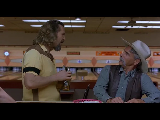 Seated at the bowling alley bar, the Stranger looks up at the Dude who is beginning to walk away, beers in hand.