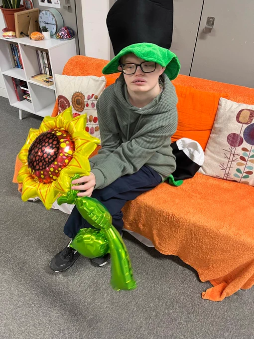 A young man wearing a green hoodie and a hat shaped like a pint of Guinness sits on an orange sofa. He's holding a foil balloon shaped like a sunflower. 