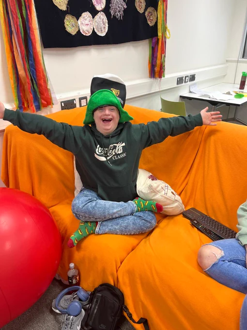 A young man sits cross legged on a sofa covered with an orange throw. He is wearing a green coca-cola hoodie, blue jeans and a hat shaped like a pint of Guinness. 