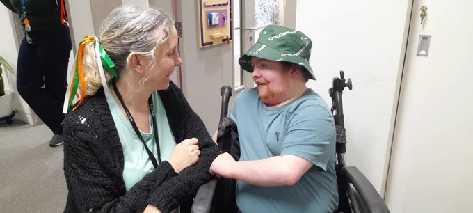 A young man sits in a wheelchair. He's wearing a teal t-shirt and a dark green bucket hat. Next to him, a lady kneels down and faces him. She has her hair in a ponytail, tied with orange, green and white ribbon in a bow. 