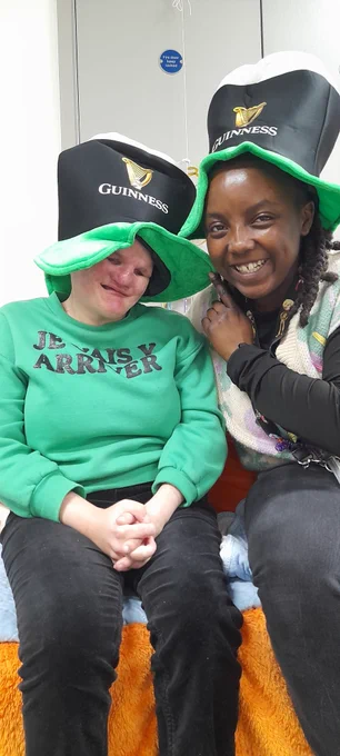 A woman wearing a bright green sweatshirt sits next to another woman. They're both wearing hats shaped like pints of Guinness and are smiling to camera. 