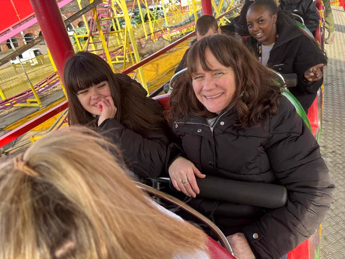 Two women both wrapped up in thick black coats are sitting in the carriage of a rollercoaster.