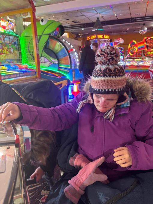 A woman wrapped up for winter in a purple coat, woolly hat and gloves feeds a 2pence into a machine at an arcade. 