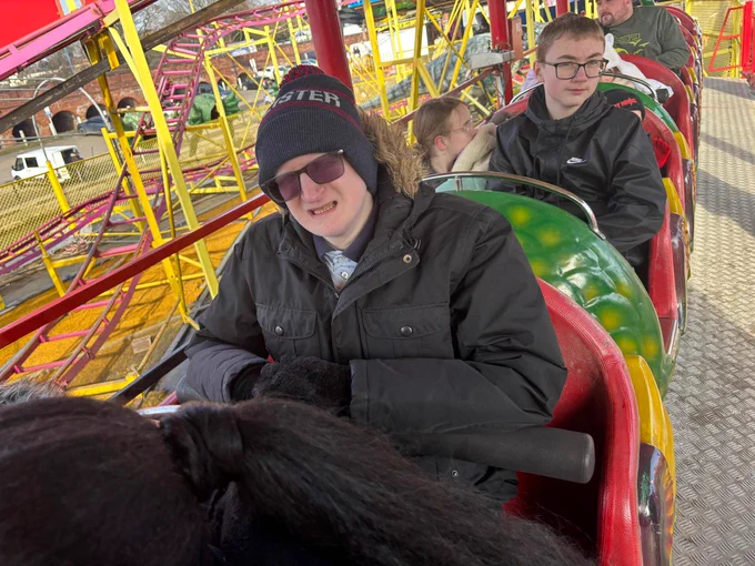 A young man wearing a coat, woolly hat and dark glasses riding a green, red and yellow rollercoaster. 