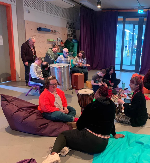 A music session taking place in a large room. There are 11 people spready out across the room, some sitting on chairs, some on bean bags, some standing. There are musical instruments including drums scattered around for people to explore. In the middle of the room is a lady crossed legged, holding a saxophone. 