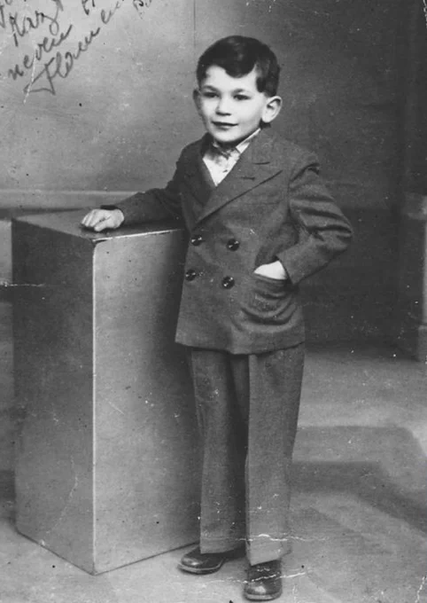 Vintage black-and-white photo of a young child in a formal double-breasted suit, standing next to a pedestal.
