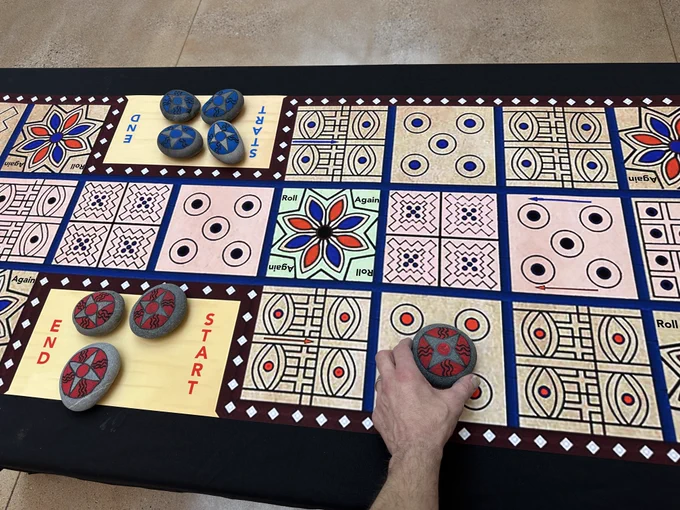 Photograph of a colorful ancient board game shown with someone's hand moving a piece made of rock