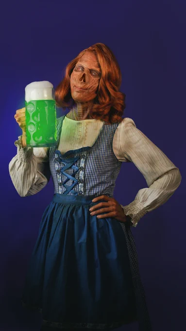A photo showing a ghoul cosplay wearing a traditional German dress holding a glass tankard of glowing liquid.
