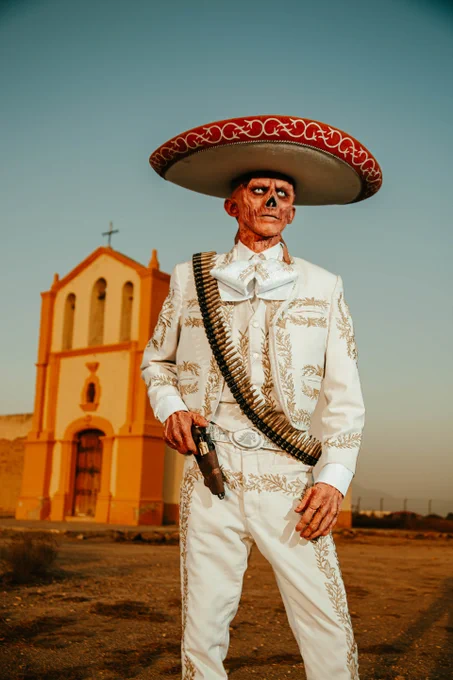 A photo showing a ghoul cosplay wearing a white mariachi outfit and sombrero. They have a bandolier over their shoulder and a revolver in a holster. In the background is a church ruin in the middle of the desert.