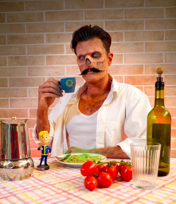 A photo showing a ghoul cosplay sat at a table eating spaghetti and holding a blue Vault 76 branded espresso cup. There is olive oil and a Vault Boy bobblehead on the table.
