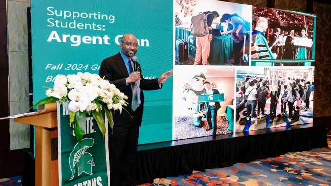 Titus Awokuse stands near podium holding a mic, giving a presentation with a large projector screen displaying events from MSU.