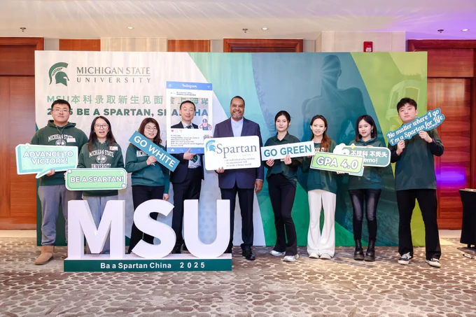 Several students and two people from MSU hold various signs as they stand in front of a large backdrop with life size MSU letters in front.