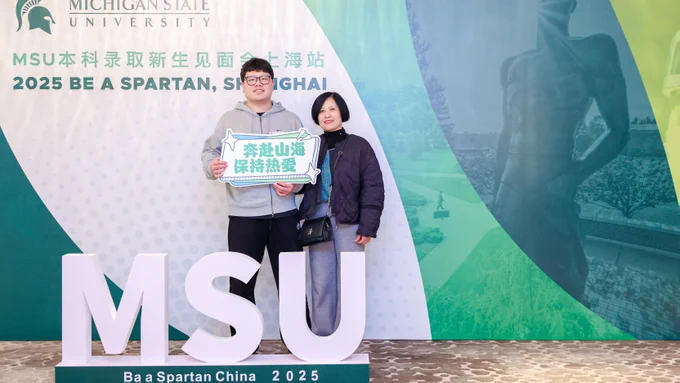 A student with parent stand in from of MSU backdrop. Student holds sign displaying Chinese characters.