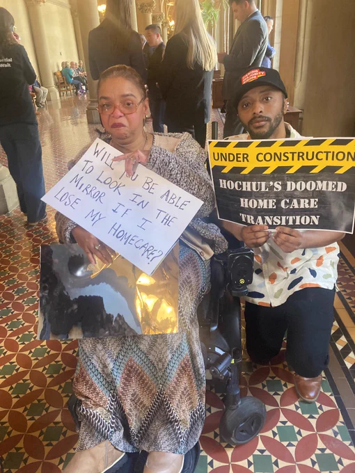Agnes Mccray, a wheelchair user and woman of color, holds a mirror and a sign that reads "Will you be able to look in the. mirror if I lose my home care?" her son holds a sign that reads "Under Construction: Hochul's DOOMED home care transition" 