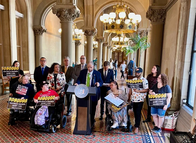 A crowd of legislators and wheelchair users with signs that read "UNDER CONSTRUCTION: Hochul's doomed home care transition" gather around a podium in the NYS capitol  