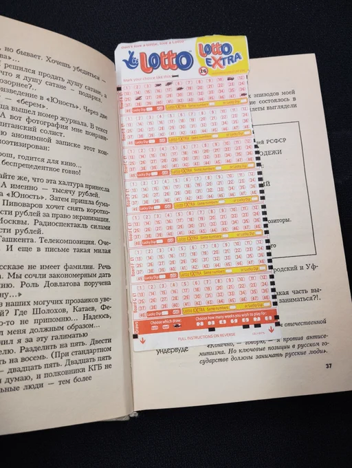 An open library book with a National Lottery ticket being used as a bookmark. The ticket is rectangular with mainly red text on a white background. It says "Lotto" and "Lotto Extra" at the top. Some lottery numbers have been selected in pen.  