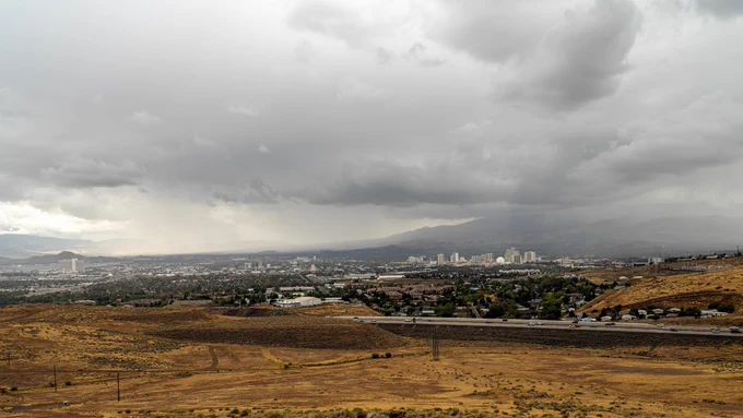 Dark storm clouds gather over Reno, Nevada, with the city's skyline visible in the distance. The foreground features dry, brown desert terrain and a busy highway, while rain showers can be seen falling over the mountains in the background, signaling wet weather ahead.