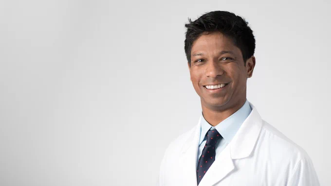 Man with short black hair smiling, wearing a shirt and tie with a white lab coat, standing in front of a light gray background.