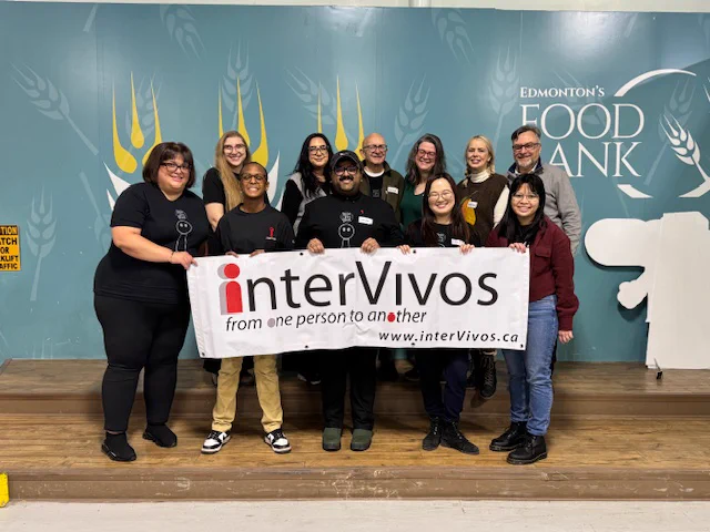 A group of interVivos board members and friends stand shoulder to shoulder on a wooden platform, holding a large banner that prominently displays the word "interVivos." The banner also includes the phrase "from one person to another" and a website address. The background features a teal-coloured wall adorned with a wheat-like design in the same colour scheme, along with white cutouts resembling animal antlers or similar shapes. Several individuals are dressed in dark-coloured clothing, including interVivos t-shirts, pants, or leggings, while others wear sweaters or jackets in shades of brown, maroon, and other colours. Some are wearing winter-style shoes. The group appears smiling and engaged, with their faces clearly visible. A yellow safety sign is present near the left side of the image. The floor is made of light brown wood.