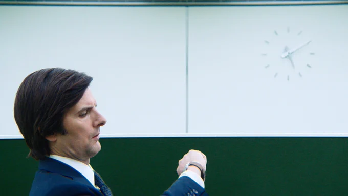 From Severance. Mark portrayed by Adam Scott is in an office at Lumon looking down at his watch. His side profile is visible. He is wearing a suit and there is a clock on the upper right side of the room.
