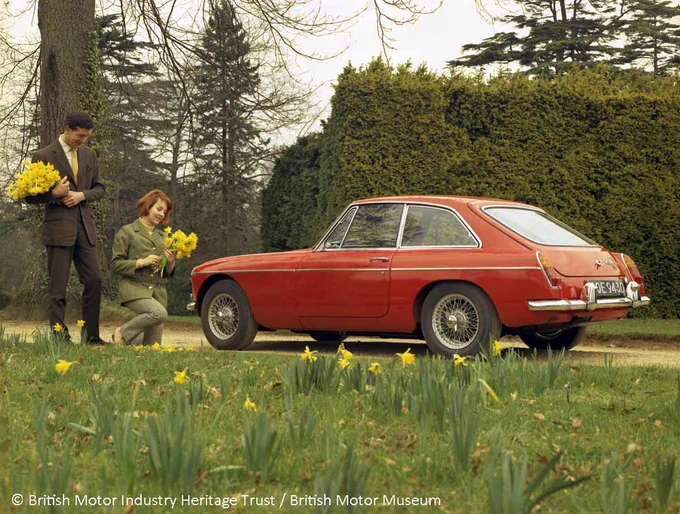 A colour photograph of a red MGB GT and a man and a woman holding bouquets of daffodils. The man is standing behind the lady who is kneeling in front of the car, which is facing towards them and parked on a road by some grass and more daffodils. 
