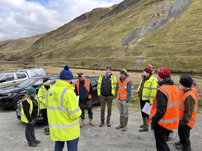 Colleagues stood talking while wearing high-vis PPE in a car park with grass covered hills in the background