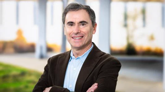 Portrait of Daniel Haskel, smiling, wearing a brown jacket with a light blue collared shirt, standing with arms crossed in an outdoor environment.