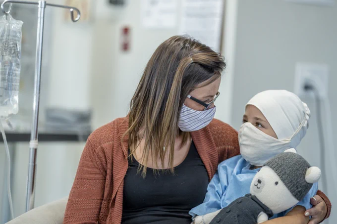 "A mother hugs her young daughter while she receives cancer treatment. Both are masked, and the child holds a plush toy"
