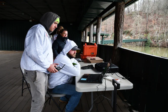 Two men in sweatshirts and a woman work at a table with a computer and other technical equipment, while looking out onto a lake. (Photo Credit:  Nayelli Guerrero, U.S. Army Corps of Engineers, Wilmington District; modified by the Army Geospatial Center) 
