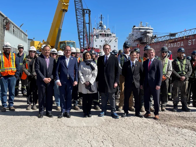 A group, including Minister Ehassai and Minister Anand, pose for a photo in front of a ship at the shipyard. 