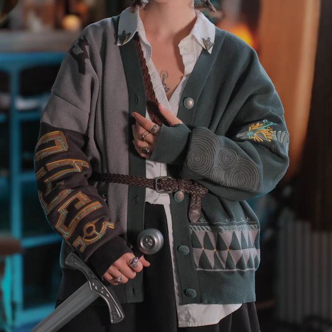 Person wearing a teal, grey, gold and cyan designed cardigan with video game inspired design, a dragon, archaic style, embroidered and jacquard knit, made by knock thrice. Person is holding a sword and wearing  rings